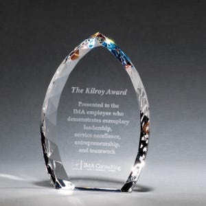 May include: A clear, teardrop-shaped crystal award with faceted edges. The front is etched with the text: The Kilroy Award, Presented to the IMA employee who demonstrates exemplary leadership, service excellence, entrepreneurship, and teamwork, IMA Consulting.