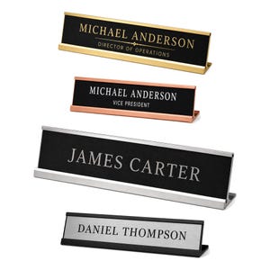 May include: Four desk nameplates with metal frames and black or silver inserts. The top plate is gold with black text reading Michael Anderson Director of Operations. The second is copper with white text reading Michael Anderson Vice President. The third is silver with James Carter. The bottom is black with Daniel Thompson.