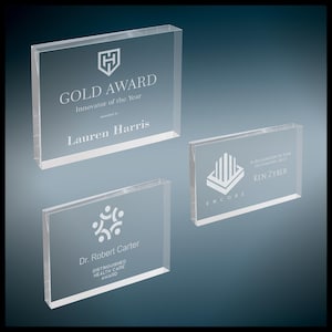 May include: Three rectangular clear acrylic awards with etched white text and logos. The top award reads GOLD AWARD Innovator of the Year awarded to Lauren Harris. The right award reads ENCORE IN RECOGNITION OF YOUR OUTSTANDING SKILLS KEN ZYBER. The bottom award reads Dr. Robert Carter DISTINGUISHED HEALTH CARE AWARD.