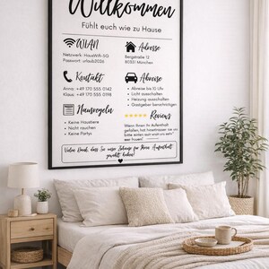 May include: A framed sign with German text, "Willkommen," above information about Wi-Fi, contact details, and house rules. The sign is above a bed with beige bedding, pillows, and a wooden tray with a mug.