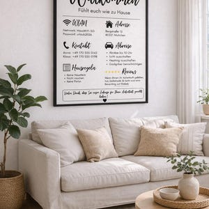 May include: A framed sign with the German word "Willkommen" in elegant script, displayed above a beige sofa with decorative pillows. The sign includes contact information, house rules, and Wi-Fi details. A small plant and a coffee table with a tray complete the scene.