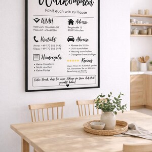 May include: A framed sign with German text, including "Willkommen" and house rules, hangs above a wooden table set for dining. The sign provides Wi-Fi details and contact information. A vase with greenery sits on the table.