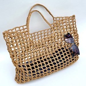 May include: A tan woven straw tote bag with an open grid pattern and two top handles. Inside the bag, a dark blue patterned fabric item is visible, and a pair of black-framed sunglasses with dark lenses is resting on the front of the bag.