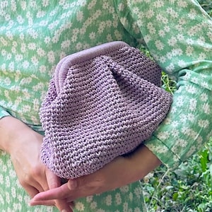 May include: A woman wearing a green floral dress and sunglasses holds a lavender crocheted clutch purse.