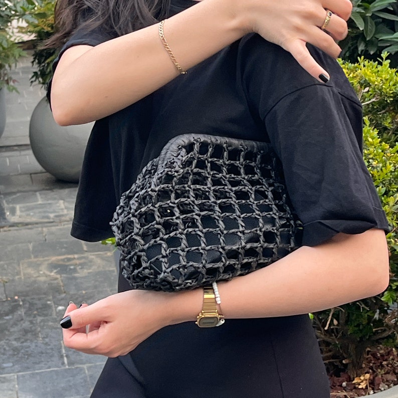 May include: A black clutch bag featuring a structured frame and an exterior layer of thick, woven black netting. The bag has a rounded, pouch-like silhouette with a solid black interior lining visible through the open grid pattern of the outer shell.