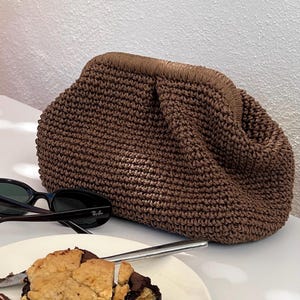 May include: A brown, woven clutch bag with a gathered top. The bag is made of a textured material and has a rounded shape. Black sunglasses and a plate with a dessert are in the foreground.