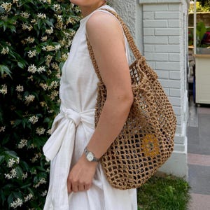 May include: A tan, open-weave crochet tote bag with a diamond pattern and two shoulder straps. The bag is constructed from a natural-colored fiber material and features a visible, circular yellow sunflower-like detail inside the main compartment.