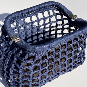 May include: A navy blue handbag featuring a structured, open-top design with a grid-like, woven mesh construction. The bag includes a reinforced, wrapped top rim and silver-toned metal hinges at the corners, revealing a dark interior lining.