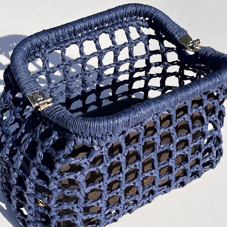 May include: A navy blue handbag featuring a structured, open-top design with a grid-like, woven mesh construction. The bag includes a reinforced, wrapped top rim and silver-toned metal hinges at the corners, revealing a dark interior lining.