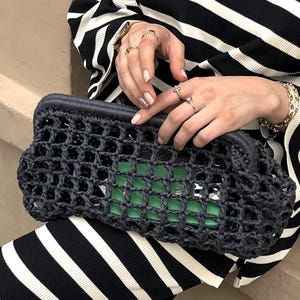 May include: A dark charcoal gray crochet clutch bag with an open-weave grid pattern and a solid frame closure. The bag is held horizontally, revealing a bright green rectangular object tucked inside the open mesh structure of the dark fiber material.