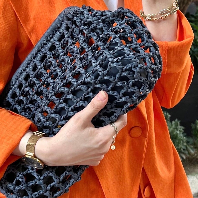 May include: A dark charcoal gray woven clutch bag featuring an open, grid-like lattice pattern. The rectangular accessory is constructed from a textured, fibrous material and is held horizontally, revealing a solid dark interior lining within the structure.