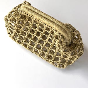 Puede incluir: Un bolso de mano beige confeccionado con material tejido similar a la rafia, que presenta un patr&oacute;n de celos&iacute;a abierto en todo el cuerpo y un marco superior estructurado y tejido s&oacute;lidamente que sirve de cierre para este accesorio alargado.