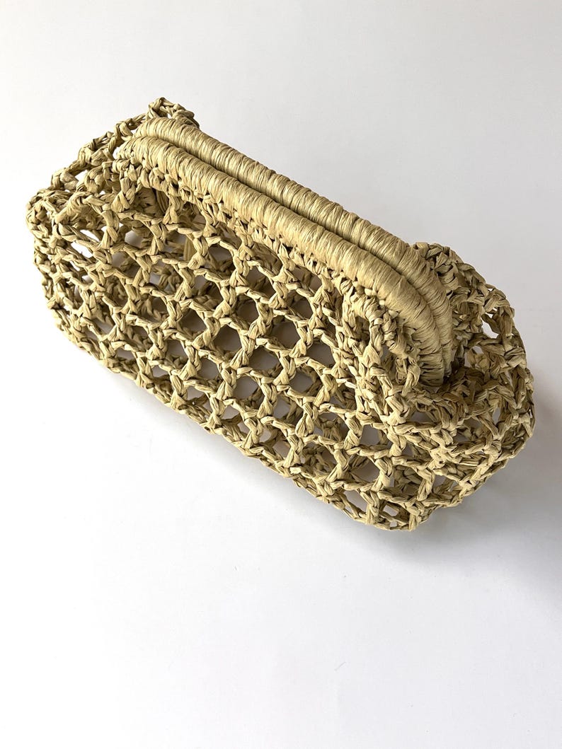 Puede incluir: Un bolso de mano beige confeccionado con material tejido similar a la rafia, que presenta un patr&oacute;n de celos&iacute;a abierto en todo el cuerpo y un marco superior estructurado y tejido s&oacute;lidamente que sirve de cierre para este accesorio alargado.