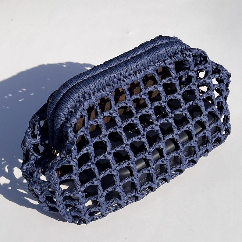 Puede incluir: Un bolso de mano azul oscuro con una estructura r&iacute;gida y un dise&ntilde;o de red tejida con cuadr&iacute;cula abierta. El exterior est&aacute; hecho de un material texturizado similar a una cuerda y contiene un objeto rectangular oscuro visible a trav&eacute;s de la malla.