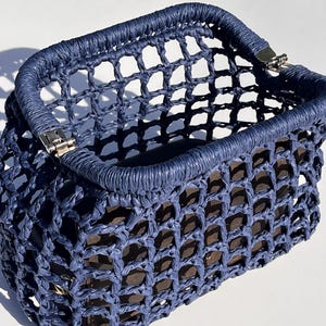Puede incluir: Un bolso de mano rectangular de color azul marino con una estructura de red de tejido abierto hecha de cuerda trenzada. El bolso tiene un marco superior r&iacute;gido y envuelto con bisagras de metal plateado en las esquinas y un forro interior marr&oacute;n.