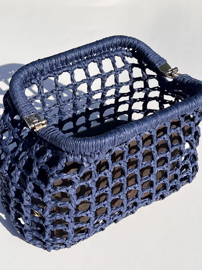 Puede incluir: Un bolso de mano rectangular de color azul marino con una estructura de red de tejido abierto hecha de cuerda trenzada. El bolso tiene un marco superior r&iacute;gido y envuelto con bisagras de metal plateado en las esquinas y un forro interior marr&oacute;n.