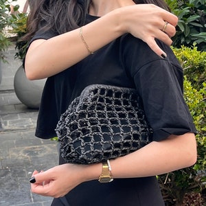 Puede incluir: Un bolso de mano tipo sobre de color negro con una textura exterior de rejilla tejida, llevado bajo el brazo. El bolso tiene una abertura de marco estructurado y un forro interior negro s&oacute;lido visible a trav&eacute;s del patr&oacute;n abierto.
