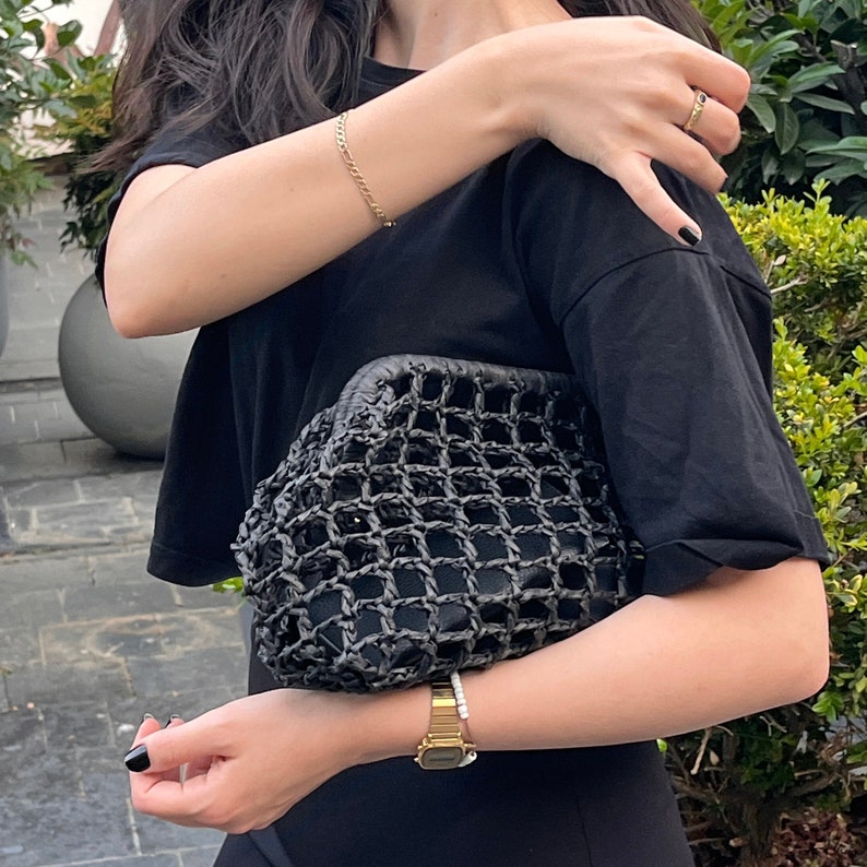 Puede incluir: Un bolso de mano tipo sobre de color negro con una textura exterior de rejilla tejida, llevado bajo el brazo. El bolso tiene una abertura de marco estructurado y un forro interior negro s&oacute;lido visible a trav&eacute;s del patr&oacute;n abierto.