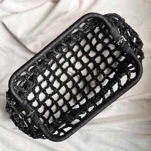Puede incluir: Una cesta rectangular negra con una estructura de red tejida y un borde s&oacute;lido envuelto. La cesta est&aacute; construida con un material fibroso oscuro, creando un patr&oacute;n de rejilla abierta en los lados y la base, con bisagras met&aacute;licas visibles.