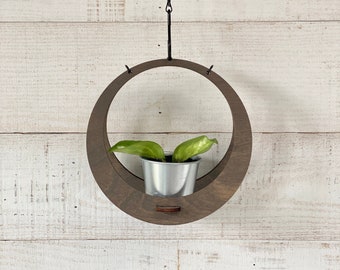 May include: A dark brown wooden hanging planter with a circular shape and a metal chain. A small green plant is growing in a silver pot inside the planter.