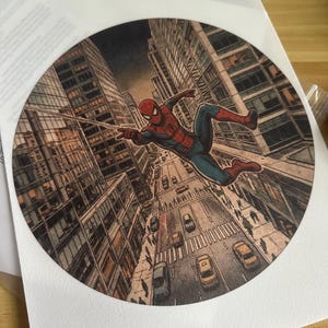 May include: A circular print featuring a comic book style illustration of Spider-Man swinging through a city street. The character is depicted in a red and blue suit, suspended above a busy urban road with cars and pedestrians between tall buildings.