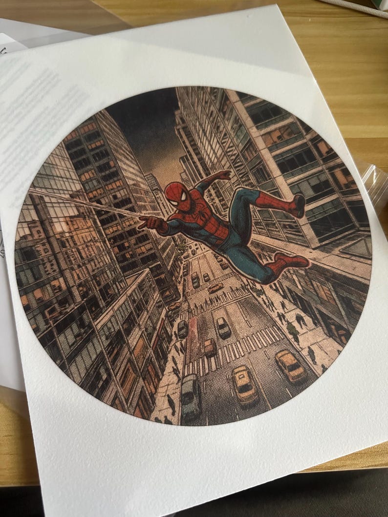 May include: A circular print featuring a comic book style illustration of Spider-Man swinging through a city street. The character is depicted in a red and blue suit, suspended above a busy urban road with cars and pedestrians between tall buildings.