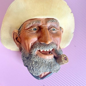 May include: A detailed, three-dimensional wall sculpture of an elderly man with a weathered face, wearing a cream-colored cowboy hat and smoking a pipe. The sculpture has a gray beard and blue eyes, with a smiling expression.