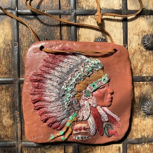 May include: A terracotta plaque with a relief sculpture of a Native American wearing a headdress with feathers. The plaque is brown with a red, white, and green headdress. The plaque has a leather cord attached to it.