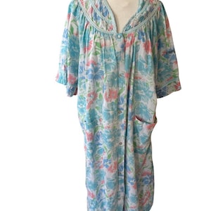 May include: A knee-length housecoat featuring a white base with a floral pattern in shades of light blue, pink, and green. The garment has short sleeves, a V-neckline with light blue trim, a full-length front button closure, and two pockets.