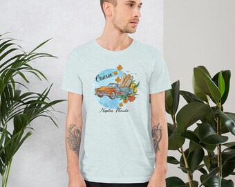 Puede incluir: Una camiseta jaspeada de color azul claro con un gráfico de un coche descapotable vintage naranja y azul que transporta tablas de surf. El diseño incluye flores de hibisco naranjas, un fondo circular azul y el texto Cruisin' Naples, Florida.