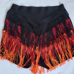 Può includere: Shorts neri con cintura nera e frange rosse, arancioni e nere.