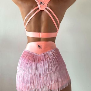 May include: A pink sports bra with a criss-cross back design and a matching pink skirt with a layered fringe detail. The bra and skirt both feature a black logo with a stylized bird and letter X.