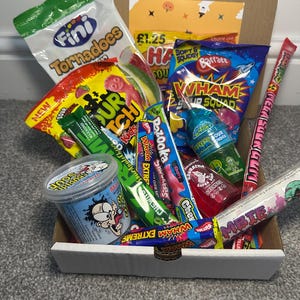 May include: A selection of sweets and candies in a cardboard box. The box includes bags of Fini Tornadoes, Wham bars, and other treats. The image shows a variety of colorful sweets, including sour candies and chewy sweets. The box is open.