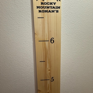 Op de afbeelding: Een houten groeimeter met de tekst "Rocky Mountain Roman's" en een bergafbeelding bovenaan. Zwarte lijnen markeren de hoogte-eenheden, met de cijfers 6 en 5 zichtbaar. De meter is gemaakt van lichtgekleurd hout.