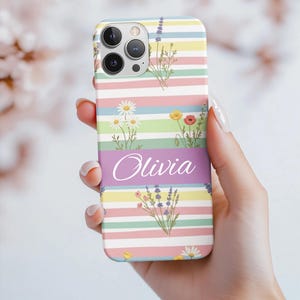 May include: A floral phone case with a pastel striped background. The case features a purple band with the name "Olivia" in cursive. Sprigs of daisies, lavender, and poppies are interspersed throughout the design. The case is held in a hand.
