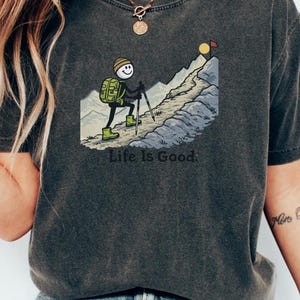 May include: Dark gray t-shirt featuring a cartoon hiker ascending a mountain, with the text "Life Is Good." The hiker has a backpack and uses trekking poles. The mountain has a sun and a flag at the peak. The shirt is paired with blue jeans.