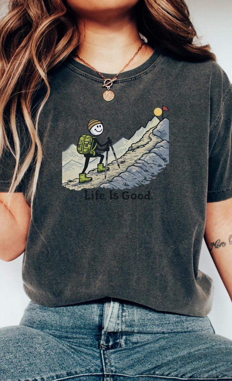 May include: Dark gray t-shirt featuring a cartoon hiker ascending a mountain, with the text "Life Is Good." The hiker has a backpack and uses trekking poles. The mountain has a sun and a flag at the peak. The shirt is paired with blue jeans.