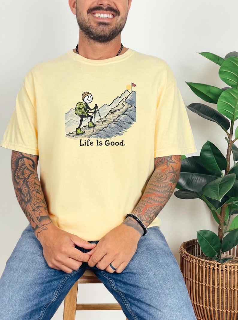 May include: Pale yellow t-shirt featuring a cartoon illustration of a hiker ascending a mountain, with the text "Life Is Good." The hiker is depicted with a backpack and hiking poles. A flag waves at the mountain's peak. The shirt has short sleeves.