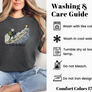 May include: Dark gray Comfort Colors t-shirt featuring a stick figure hiking up a mountain with the text "Life Is Good." The shirt also includes a washing and care guide with instructions for washing and drying. The shirt is made of Comfort Colors 1717 material.
