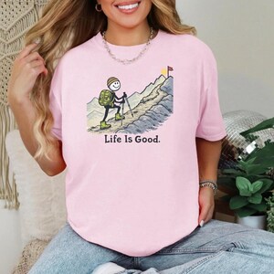 May include: A light pink t-shirt with a graphic of a stick figure hiking up a mountain, with the text "Life Is Good." The stick figure has a backpack and is using hiking poles. A yellow and red flag is at the mountain's peak.