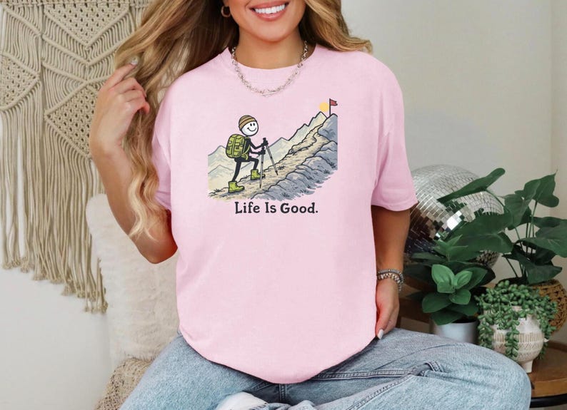 May include: A light pink t-shirt with a graphic of a stick figure hiking up a mountain, with the text "Life Is Good." The stick figure has a backpack and is using hiking poles. A yellow and red flag is at the mountain's peak.