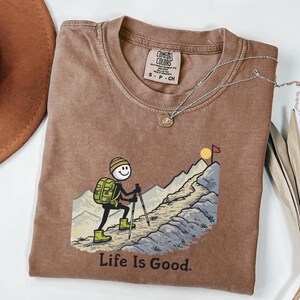 May include: A brown Comfort Colors t-shirt featuring a cartoon hiker with a backpack, climbing a mountain towards a flag. The text "Life Is Good" is printed below the graphic. A silver necklace with a pendant is draped on the shirt.