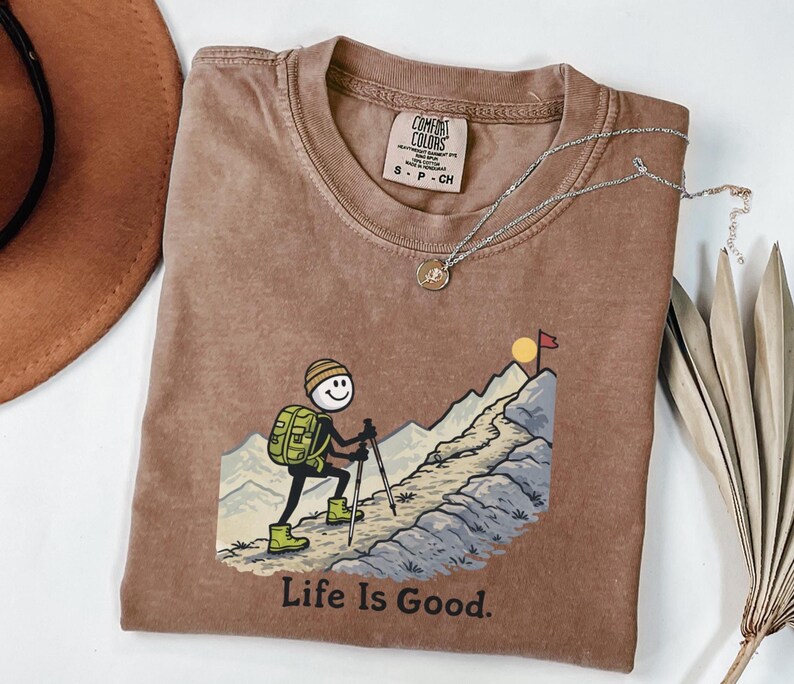 May include: A brown Comfort Colors t-shirt featuring a cartoon hiker with a backpack, climbing a mountain towards a flag. The text "Life Is Good" is printed below the graphic. A silver necklace with a pendant is draped on the shirt.