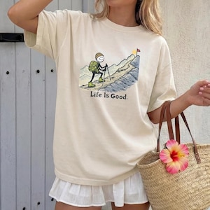 May include: Cream-colored t-shirt featuring a cartoon hiker ascending a mountain, with the text "Life Is Good." The hiker is depicted with a backpack and hiking poles. A flag waves at the mountain's peak. The shirt is paired with a white skirt and a straw tote bag.