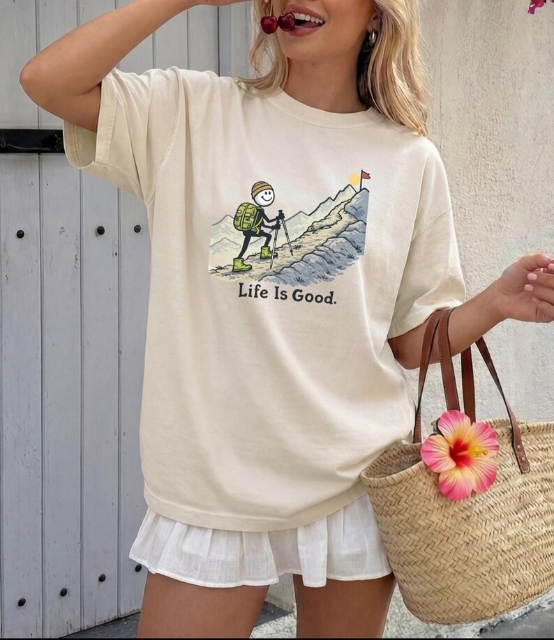 May include: Cream-colored t-shirt featuring a cartoon hiker ascending a mountain, with the text "Life Is Good." The hiker is depicted with a backpack and hiking poles. A flag waves at the mountain's peak. The shirt is paired with a white skirt and a straw tote bag.