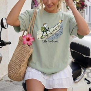 May include: Sage green t-shirt with a mountain climbing graphic and the text "Life Is Good." A woven tote bag with a pink flower hangs from the shoulder. The person is sitting on a white scooter.