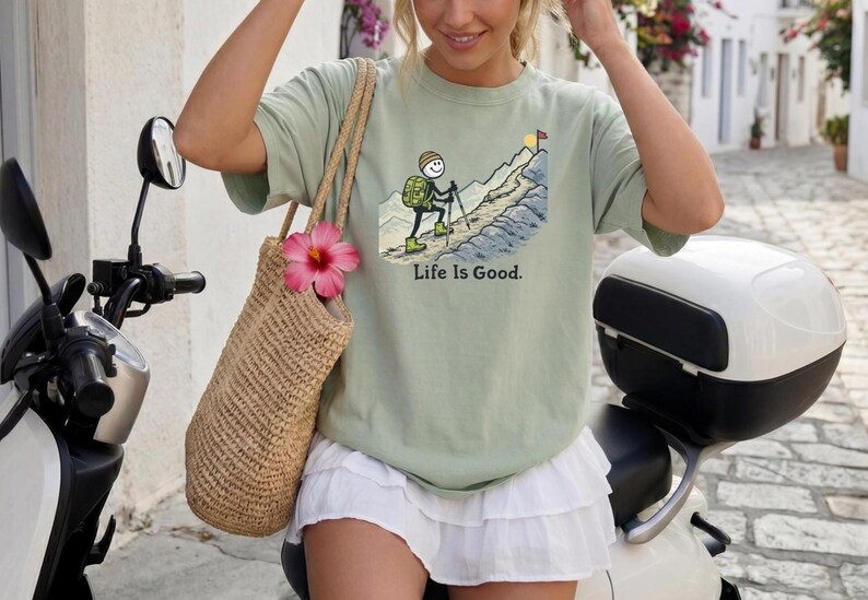 May include: Sage green t-shirt with a mountain climbing graphic and the text "Life Is Good." A woven tote bag with a pink flower hangs from the shoulder. The person is sitting on a white scooter.