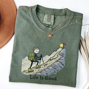 May include: Olive green Comfort Colors t-shirt with a graphic of a stick figure hiking up a mountain, the words "Life Is Good" are printed below the graphic. A gold necklace and a brown hat are also in the image.