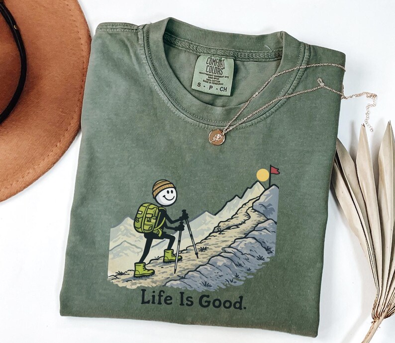 May include: Olive green Comfort Colors t-shirt with a graphic of a stick figure hiking up a mountain, the words "Life Is Good" are printed below the graphic. A gold necklace and a brown hat are also in the image.