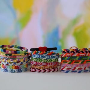 May include: Three stacks of woven friendship bracelets made from colorful embroidery floss. The bracelets feature various geometric patterns, including chevrons and stripes, in shades of blue, green, red, yellow, pink, and white, with one black bracelet featuring a red heart.